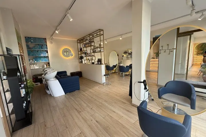La zona parrucchiere: lavatesta e divano blu, mensole in ottone con i prodotti, specchio ovale dorato e l'insegna luminosa Coco alla parete